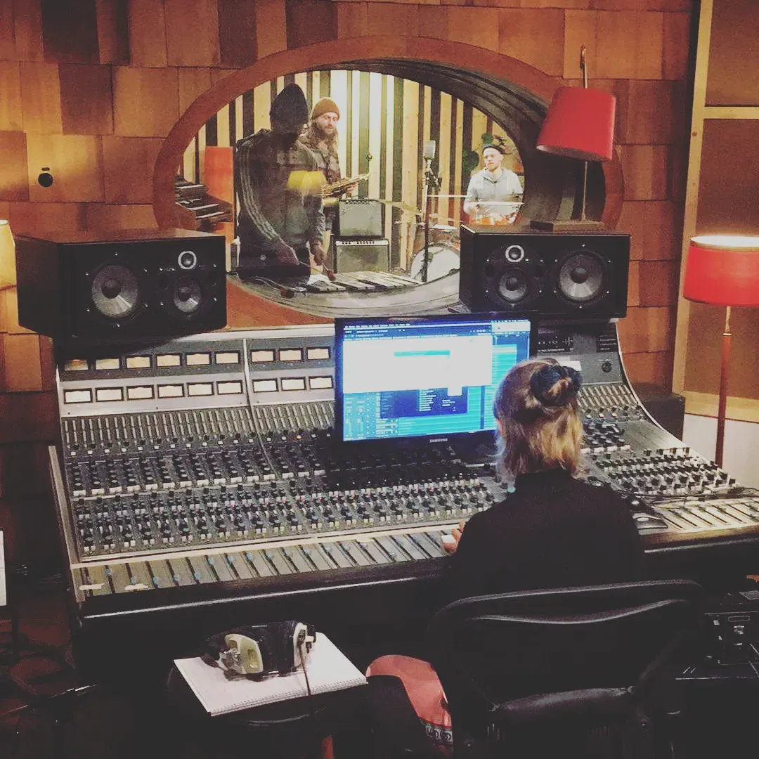Inspelning vid ett stort mixerbord, bandet spelar bakom glasrutan.